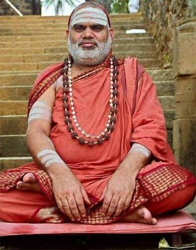 Jagadguru Sri Bharati Tirtha Mahaswamiji seated cross-legged on a mat at the foot of old stone steps, hands resting on his knees, wearing rudraksha malas and ochre robes
