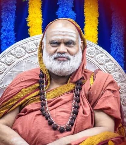Jagadguru Sri Bharati Tirtha Mahaswamiji seated on an ornate silver throne, with festive hangings of blue, yellow and red behind him