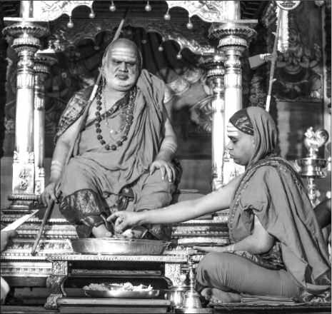 Black-and-white photograph: Jagadguru Sri Bharati Tirtha Mahaswamiji seated on an ornate throne holding the danda while a devotee seated before him performs pada puja, worship of his feet, with a plate of offerings