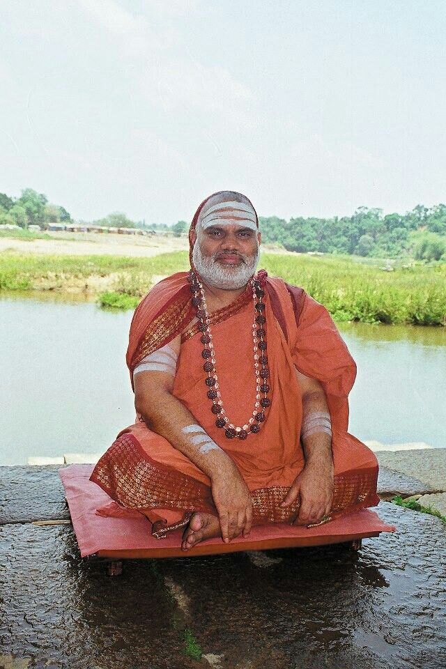 Jagadguru Sri Bharati Tirtha Mahaswamiji seated cross-legged on a raised stone platform beside a river, with paddy fields and trees on the far bank