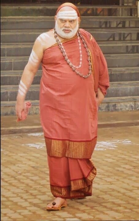 Jagadguru Sri Bharati Tirtha Mahaswamiji walking barefoot across a paved courtyard in front of wide stone steps, dressed in ochre robes with a rudraksha mala
