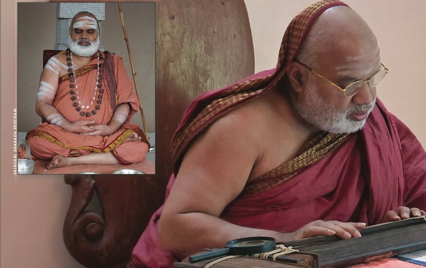 Jagadguru Sri Bharati Tirtha Mahaswamiji, wearing spectacles, reads a text laid open on a low desk; a framed photograph of the Jagadguru seated in meditation stands behind him
