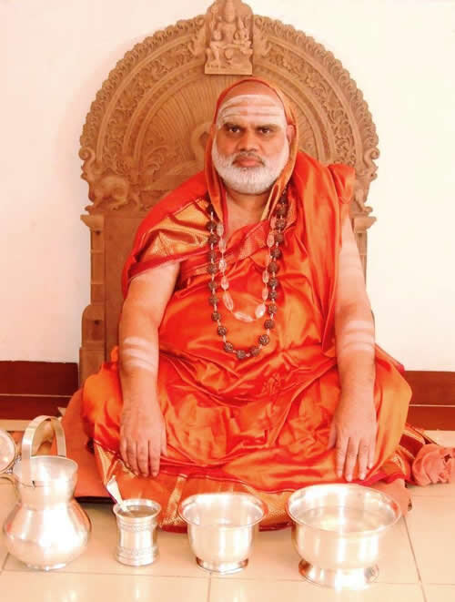 Jagadguru Sri Bharati Tirtha Mahaswamiji seated before an ornately carved wooden throne, with silver ritual vessels arranged on the floor before him