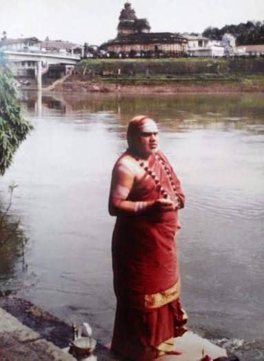 Jagadguru Sri Bharati Tirtha Mahaswamiji standing at the edge of the Tunga river; across the water the stone Vidyashankara temple rises above the far bank