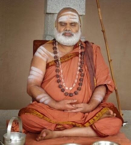 Jagadguru Sri Bharati Tirtha Mahaswamiji seated in meditation with eyes closed and hands folded in his lap, the sannyasi’s danda resting against his shoulder, silver vessels beside him